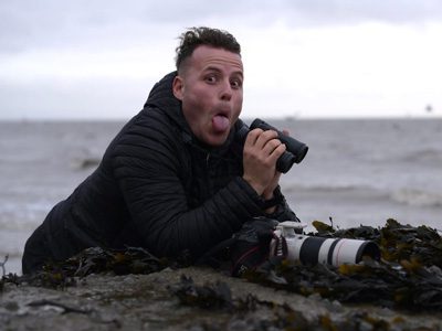 Man lying on a rocky shoreline while photographing with a camera.