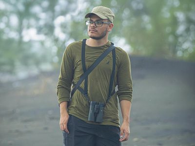 Man wearing glasses and a hat standing outdoors with binoculars around his neck.