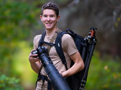 Man holding a camera and tripod over his shoulder, smiling.