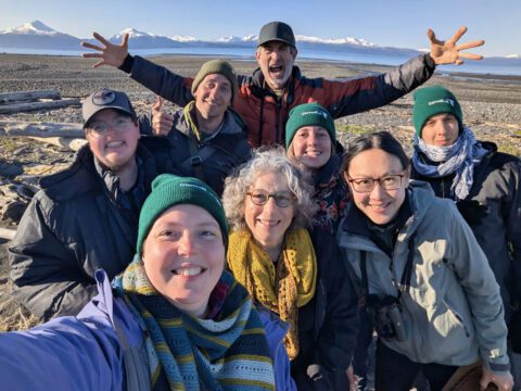 A group of eight people smile at the camera with snow-capped mountains far behind them.