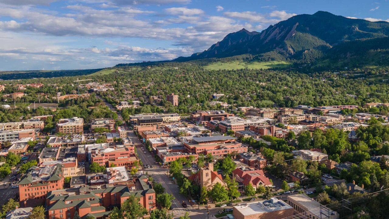 An aerial view of a town surrounded by lush greenery, with red-brick buildings and streets in the foreground. In the background, dramatic mountain peaks rise under a partly cloudy blue sky.