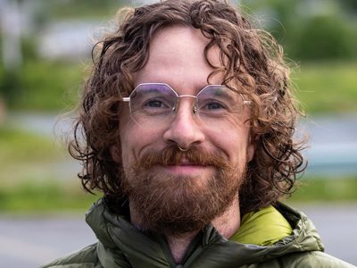 Man with curly hair and glasses smiling outdoors.