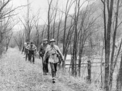 A group of people walking along a narrow forest trail beside a river, surrounded by leafless trees in a natural, serene environment.