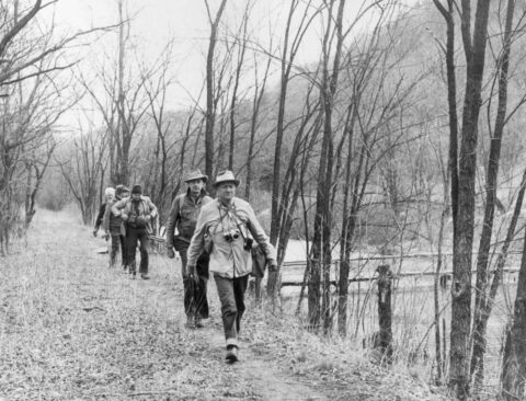 A group of people walking along a narrow forest trail beside a river, surrounded by leafless trees in a natural, serene environment.