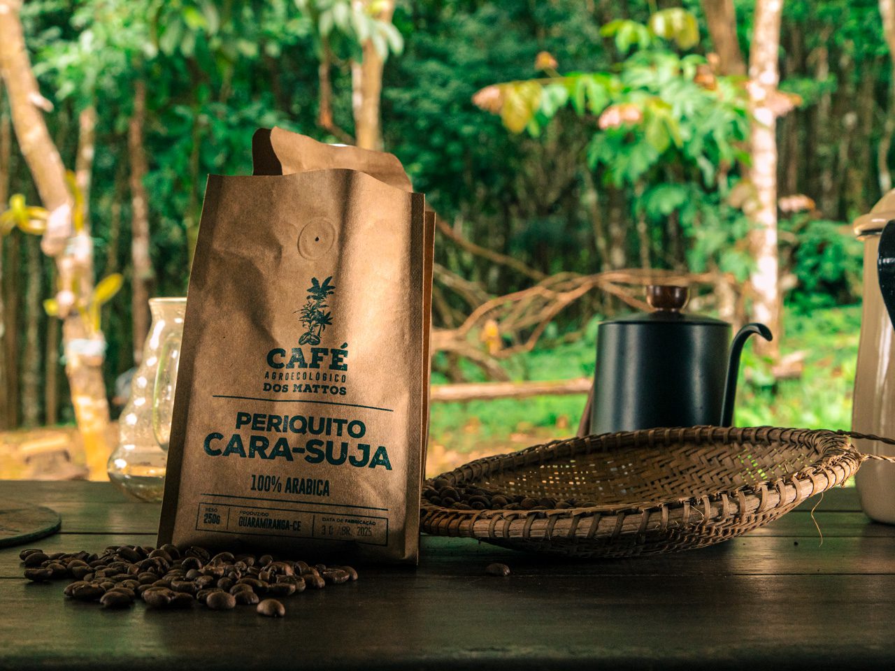 A brown paper bag labeled “Periquito Cara-Suja – 100% Arábica” sits on a wooden table beside scattered coffee beans, a woven basket, and a black kettle