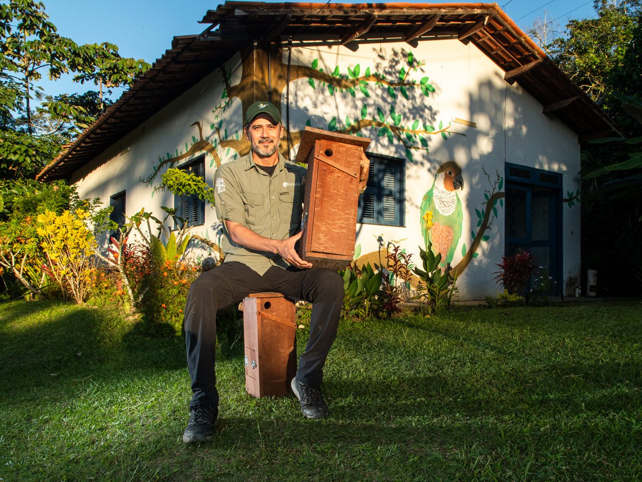 A man sitting on a stool in front of a painted building, holding a large wooden nest box on his lap.