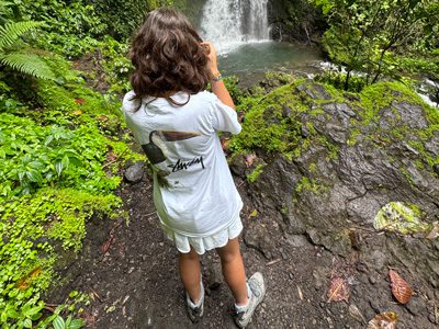 Girl standing on a forest trail photographing a waterfall, seen from behind.