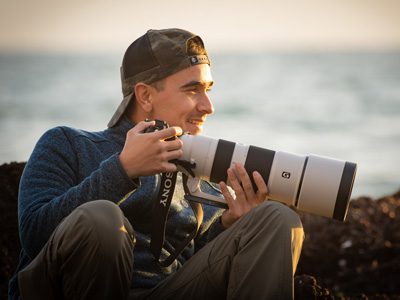 Man sitting outdoors by the water holding a large camera and smiling.