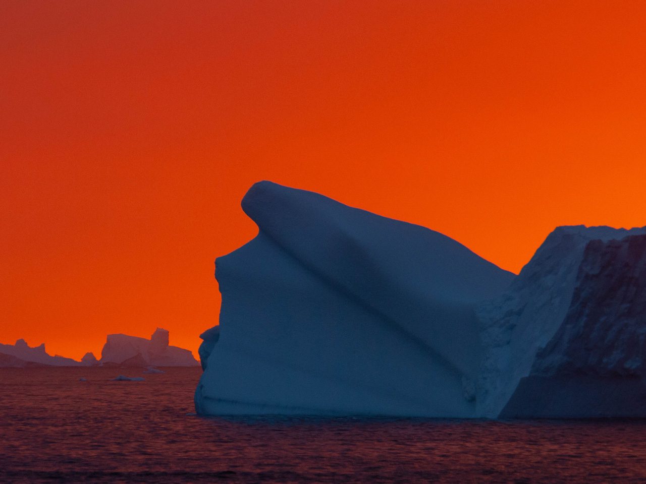 Iceberg floating on the ocean at sunset with an orange sky.