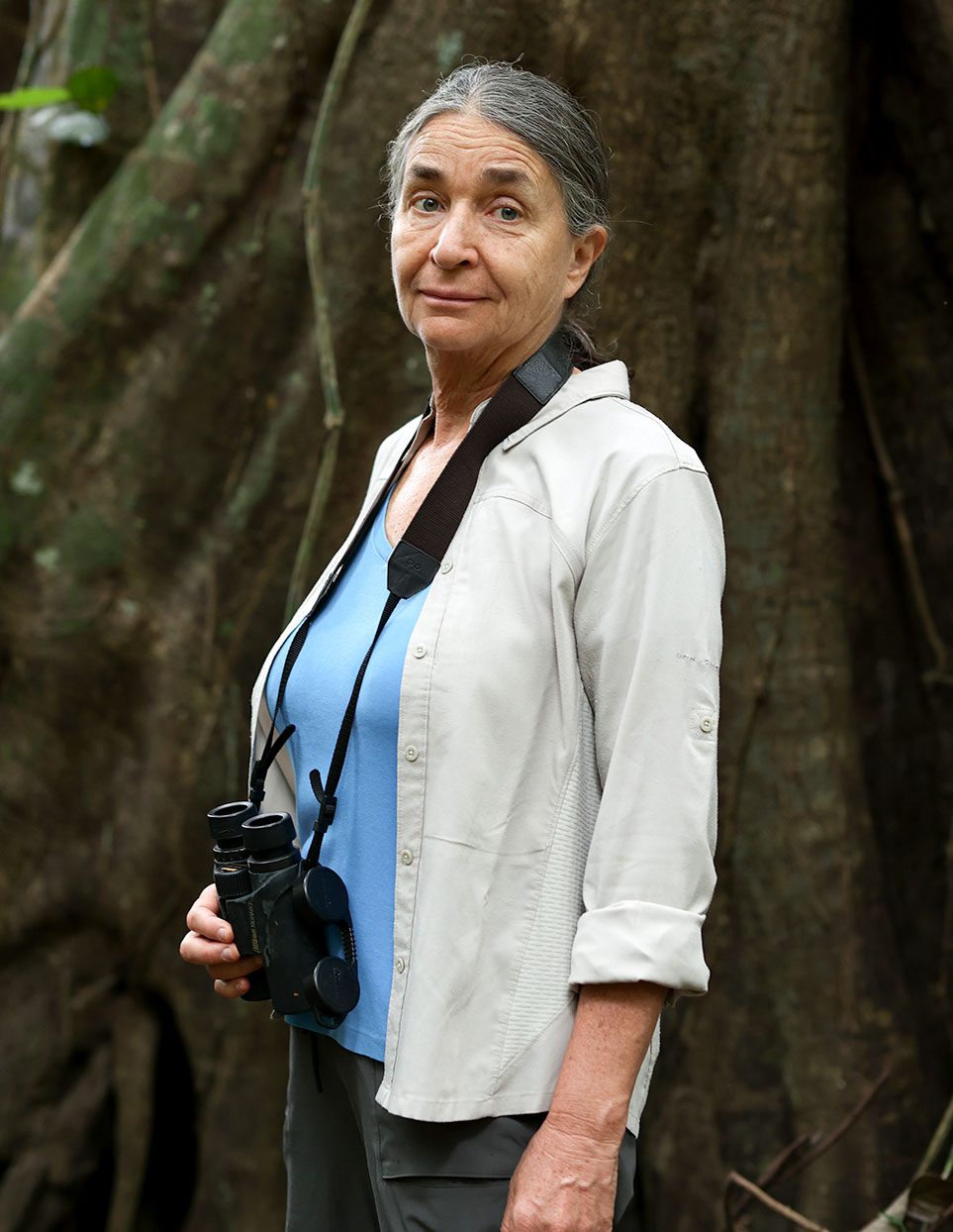 An older woman in a white shirt and binoculars around her neck, stands next to a the roots of a giant tree.