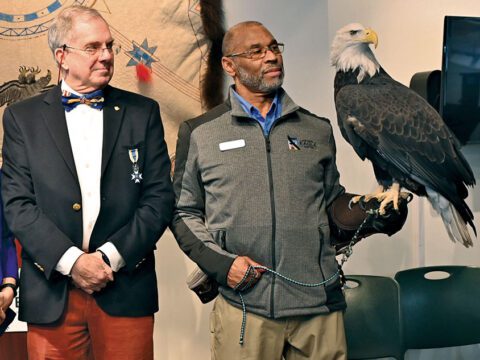 Two men stand side by side and one holds a large black bird with a white head, large yellow feet and a large, hooked bill.