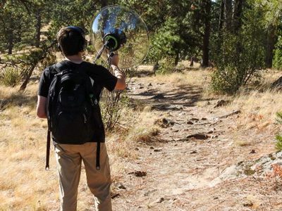 Man walking on a forest trail with a backpack and holding a parabolic microphone, seen from behind.