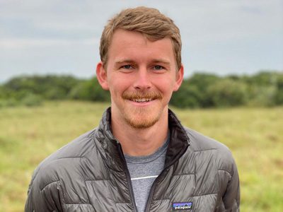 Man smiling outdoors with fields and sky in the background.