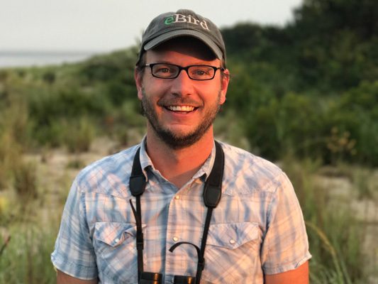 Man wearing glasses and binoculars around his neck, smiling outdoors.