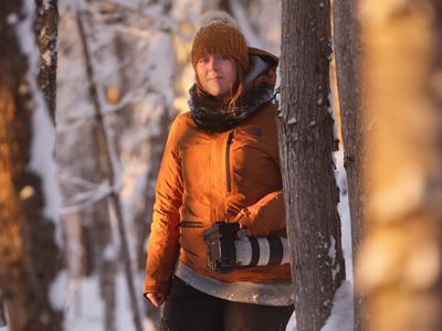 Woman in an orange jacket standing in a snowy forest with a camera hanging at her side.