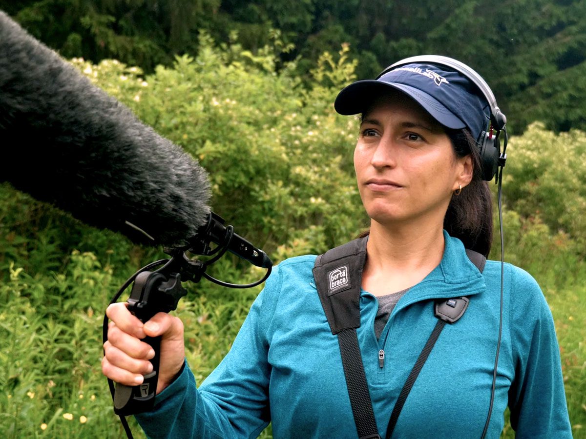 Woman in blue cap and shirt with headphones holds a fuzzy microphone.